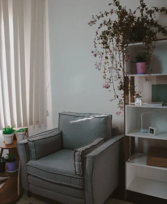 Espaço de atendimento com poltrona cinza, estante branca decorada com plantas e objetos, e iluminação natural proveniente da janela, criando um ambiente acolhedor.