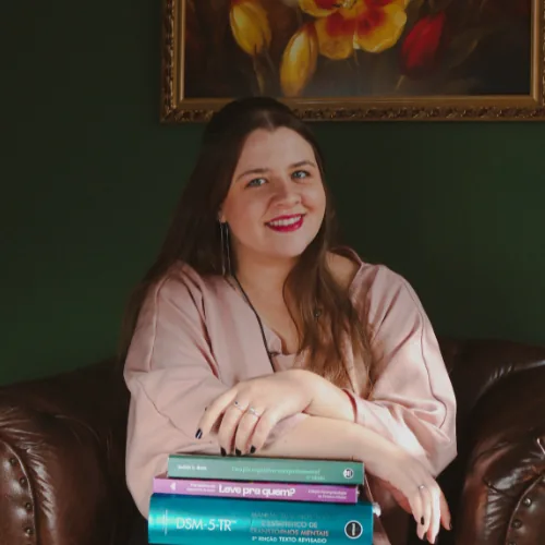 Gabriella Pankiv Kitzig, psicóloga, sentada em uma poltrona com livros de psicologia nas mãos, em um ambiente acolhedor com quadro floral ao fundo.