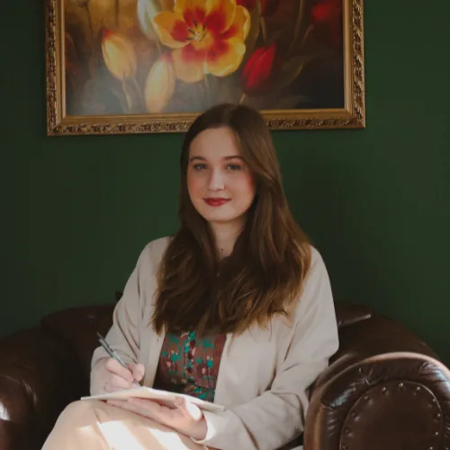 Marina Paula Zanini Sacoman, psicóloga, sentada em uma poltrona com bloco de anotações e caneta, em ambiente interno com parede verde e quadro floral ao fundo.
